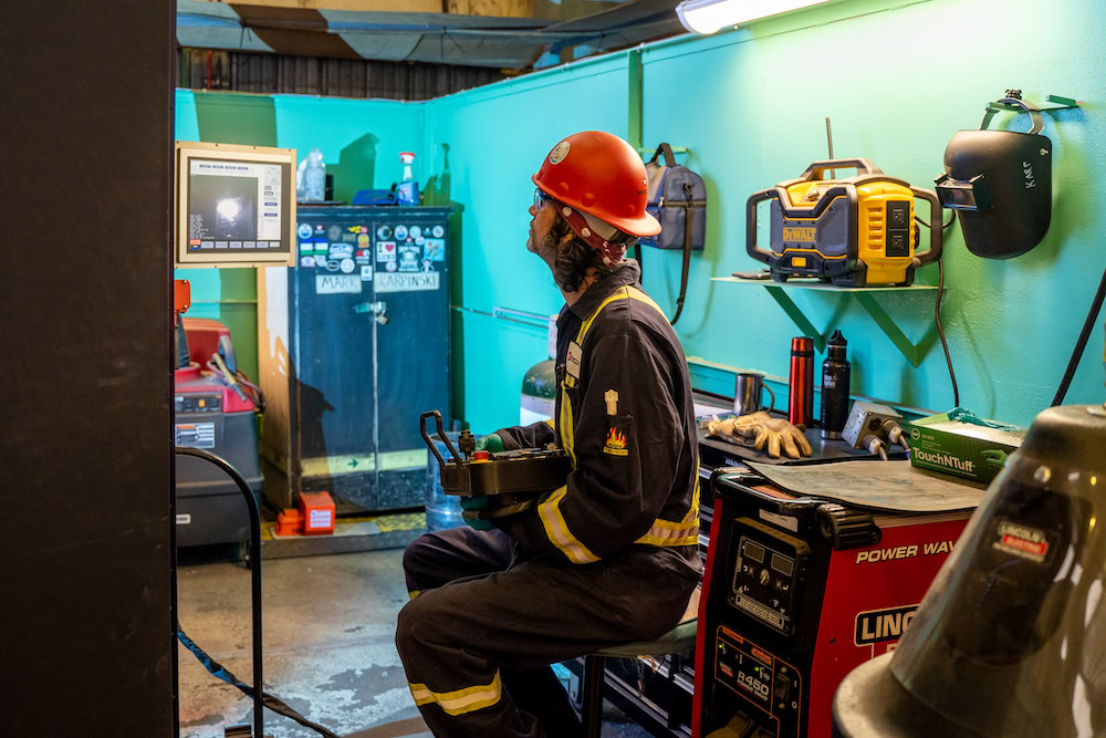 A worker in safety gear sits in a maintenance room, holding a control device. The room has teal walls and is equipped with tools, machinery, a monitor, and a welding mask hanging on the wall. A helmeted individual appears focused on a task.