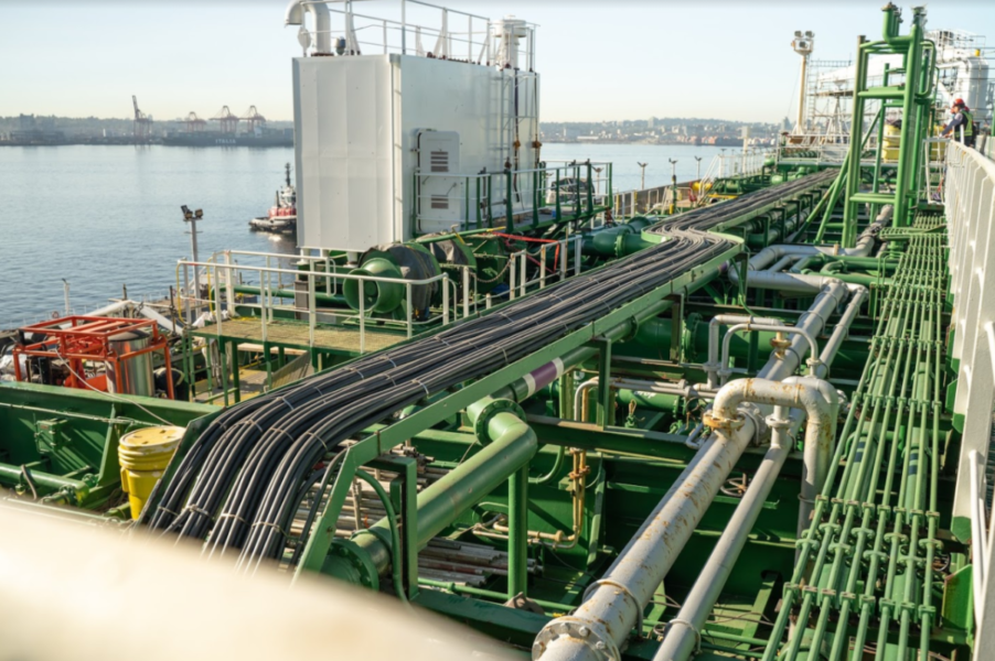 The image shows a large industrial ship deck with complex green and silver piping systems and machinery. It is docked near a shoreline, with cranes visible in the background under a clear sky.