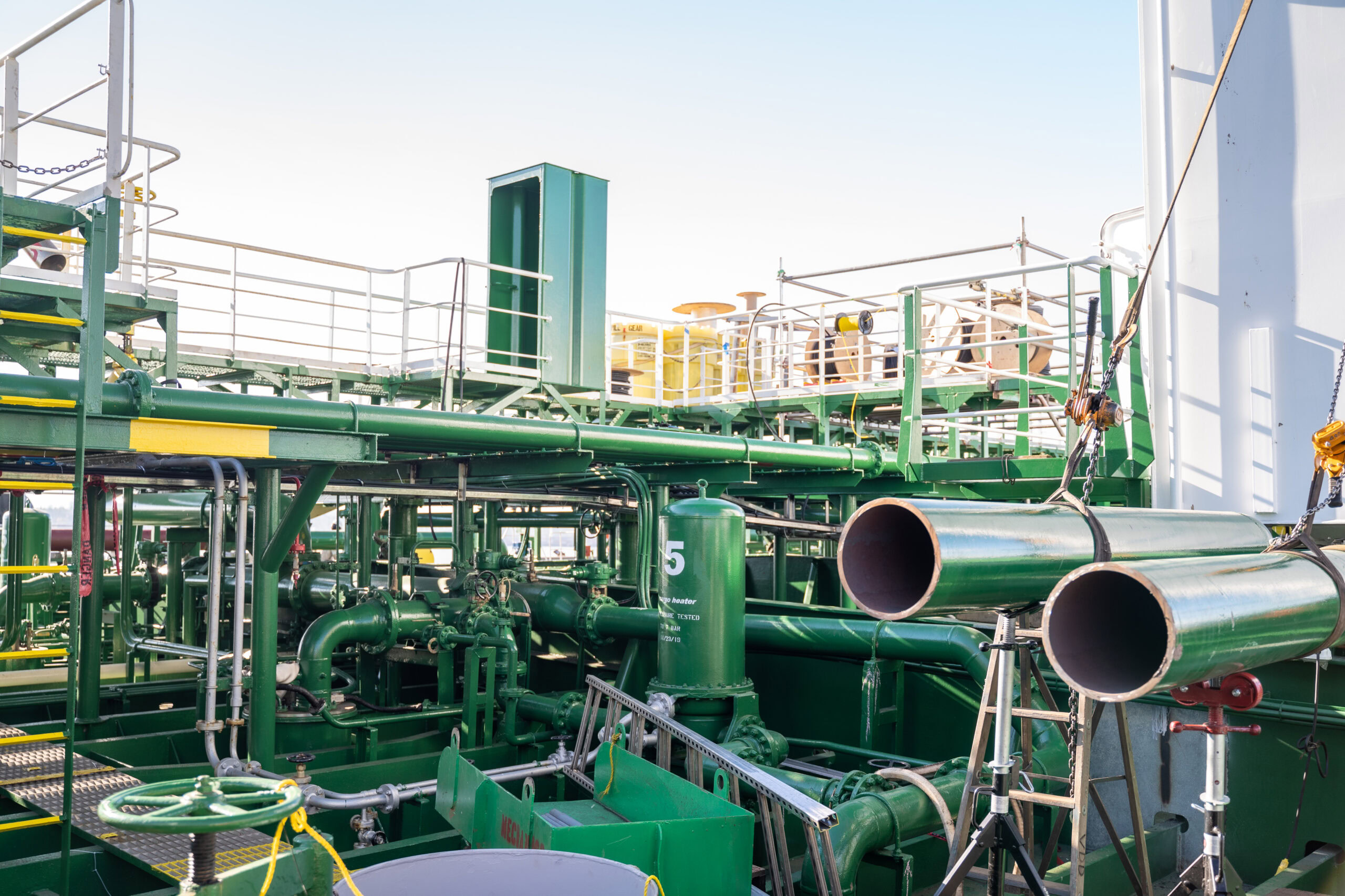 A complex industrial structure with green pipes and metal platforms. Several cylindrical pipes are stacked on a metal stand in the foreground. Safety rails and ladders are visible, along with a clear blue sky in the background.