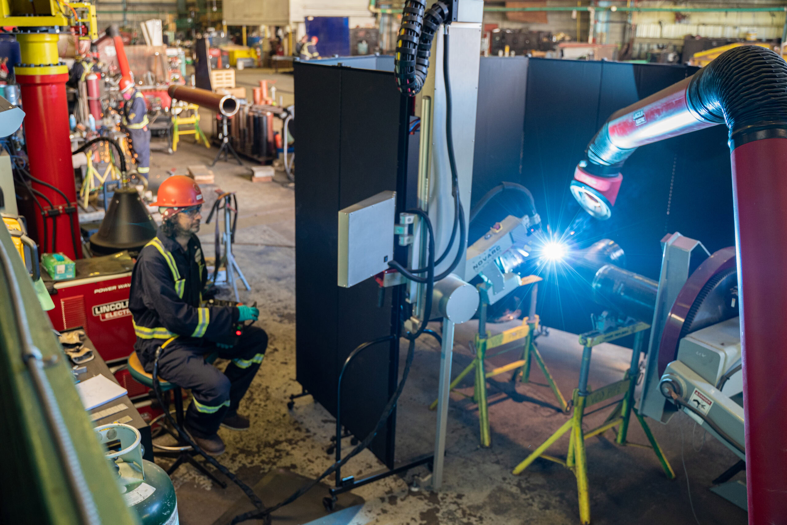 A worker in protective gear and a red helmet sits operating a large robotic welding arm in an industrial workshop. The arm emits a bright light as it works on metal tubing. The workshop is filled with various tools and equipment.