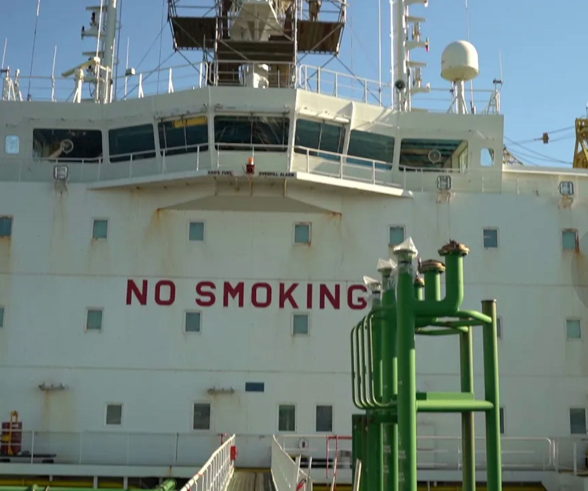 The image shows the side of a ship with the words "NO SMOKING" painted in large red letters. Various pipes and equipment are visible on the deck, with a clear blue sky in the background.