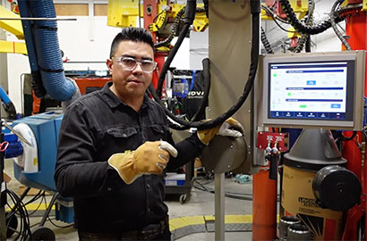 A person wearing safety glasses and gloves stands in a workshop, next to industrial equipment and a monitor displaying data. The background features various machinery and cables.