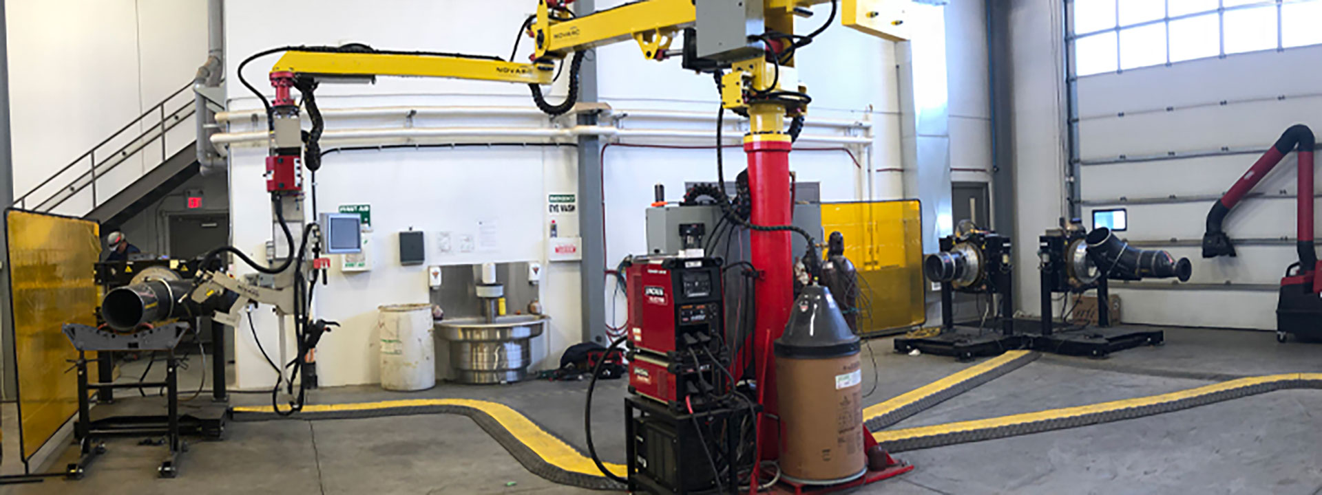 A workshop with industrial robotic machinery, including a large yellow robotic arm and welding equipment. Several control panels and safety barriers are visible.