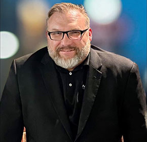 A bearded man with glasses is smiling and wearing a black suit jacket over a black shirt. The background is blurred with lights in various colors.