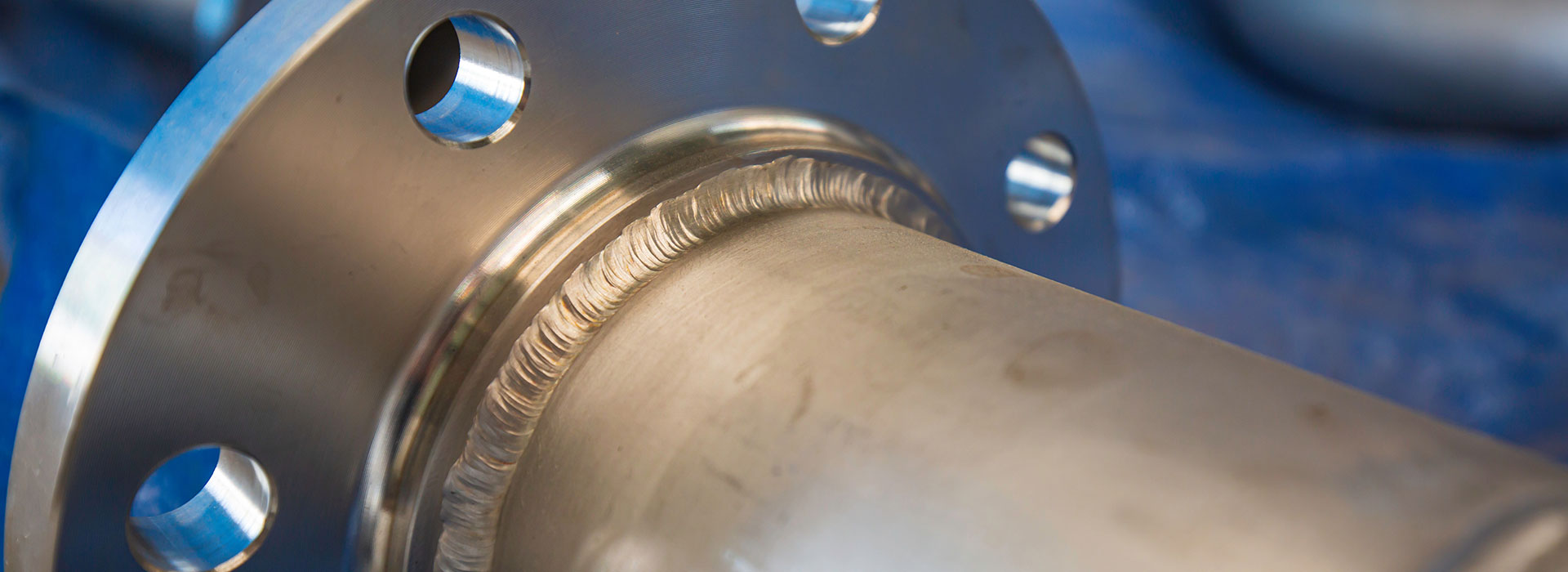 Close-up of a metallic pipe section with a smooth weld seam and flange, against a blue background. Several holes are visible in the flange for bolt fitting. The pipe surface has a polished finish.