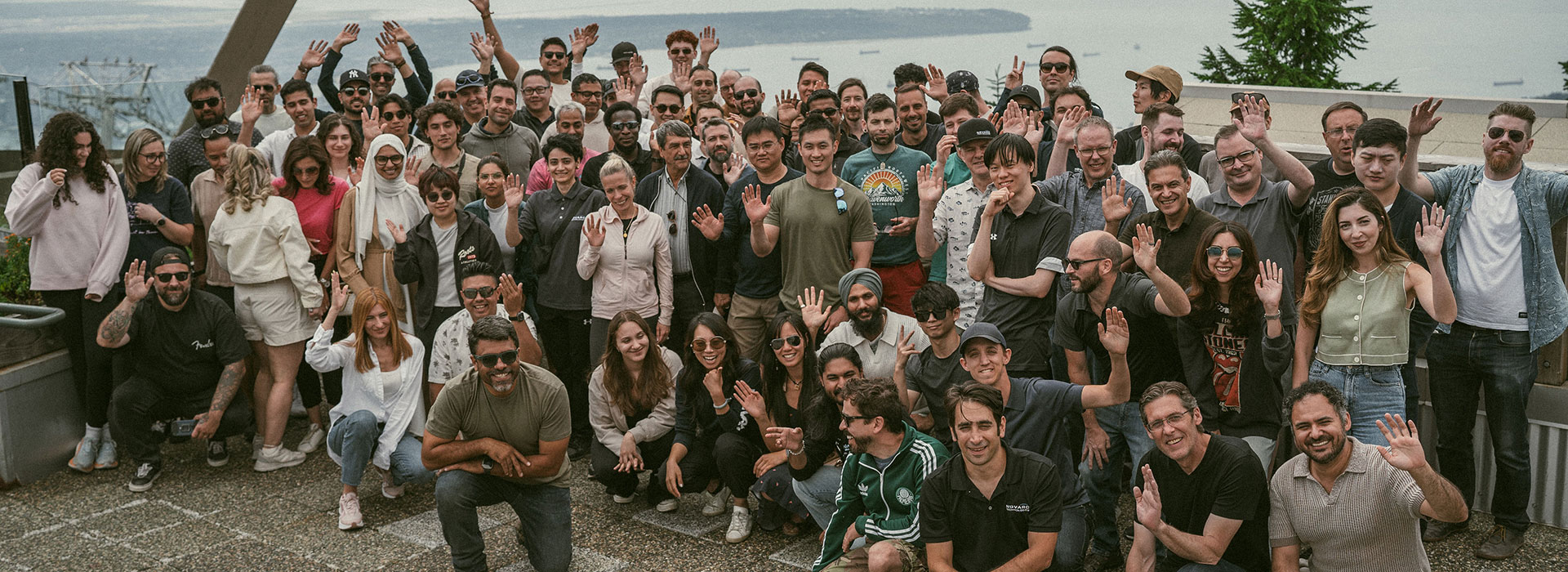 A large group of people pose for a photo on a rooftop with a distant view of the sea and ships. Some people are waving or making peace signs, and the atmosphere is cheerful. The background includes trees and a clear sky.