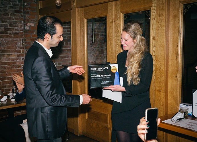 A man in a suit receives a certificate from a woman in a black outfit inside a room with wooden walls and brick accents. They are smiling, and a person in the background is applauding.