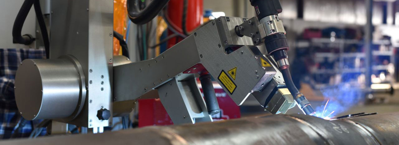 A robotic welding arm in a workshop setting welds a large metal pipe, with sparks visible. Shelves with tools and equipment are blurred in the background.