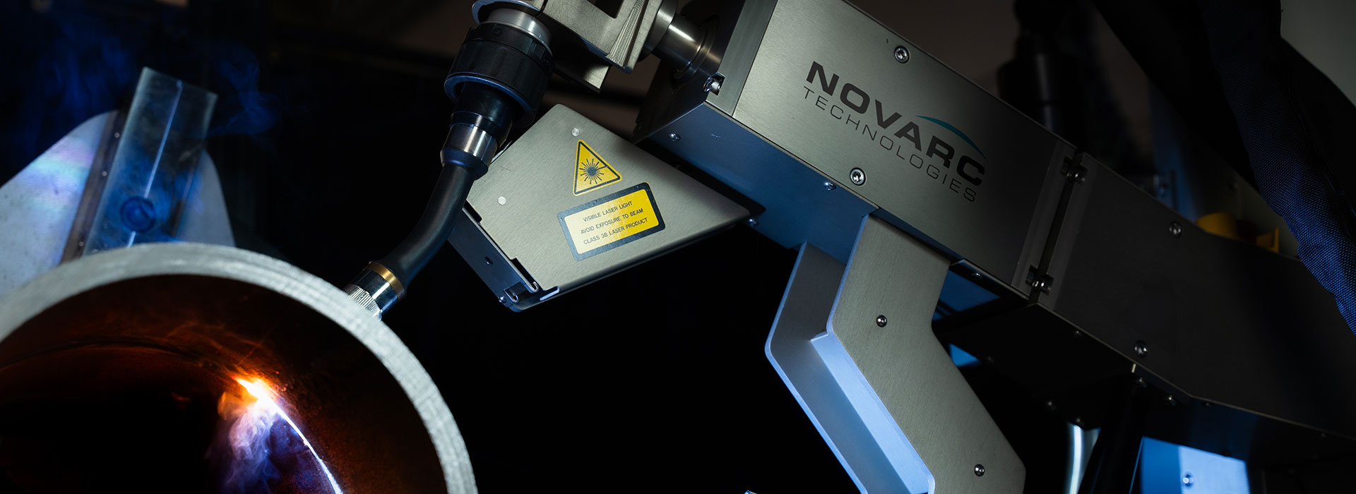 A robotic arm from Novarc Technologies is positioned over a large metal cylinder, emitting a bright welding arc. The setup appears to be part of an industrial welding process, with blue sparks visible around the metal.