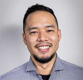 A smiling person with a beard and short hair, wearing a checkered shirt, stands against a plain background.