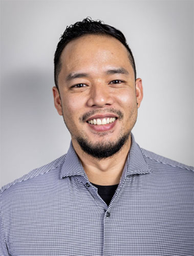 A person with short, dark hair and a beard smiles warmly at the camera. They are wearing a gray checkered shirt against a plain, light gray background.