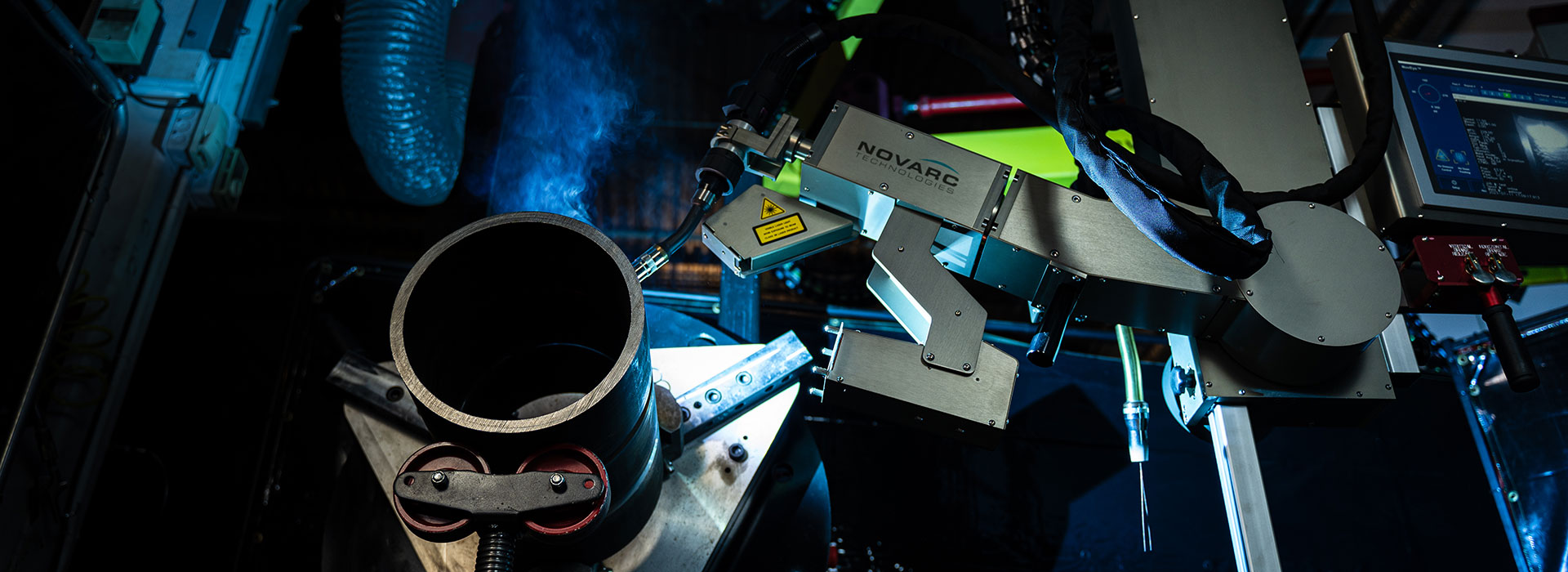 A robotic welding arm operates on a cylindrical metal piece, surrounded by industrial equipment under dim lighting. A screen is visible on the right, displaying digital data. Blue light and smoke add to the high-tech atmosphere.