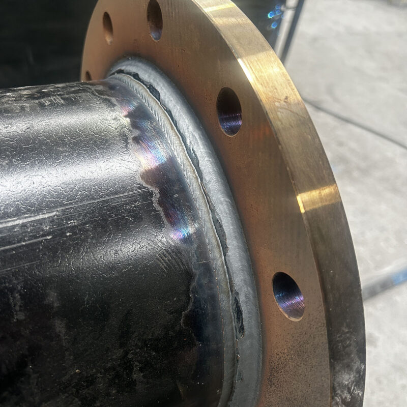 Close-up of a metallic pipe with a welded joint next to a circular flange. The weld has a slightly uneven texture with rainbow discoloration. The flange has several holes around its edge. The background is blurred concrete.