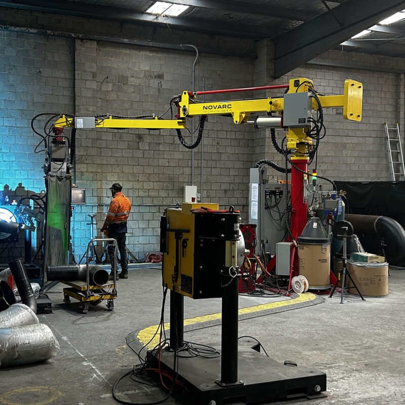 A person operates a large industrial welding robot in a factory setting. The machine, marked "Novarc," is positioned near metal pipes and equipment, with concrete walls and a high ceiling visible in the background.