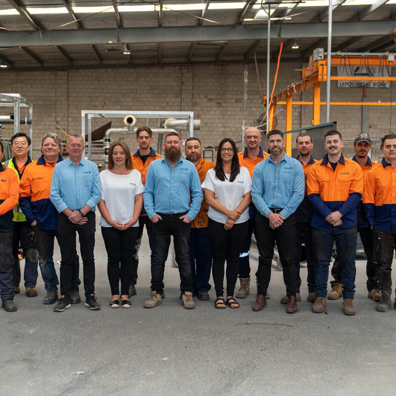 A diverse group of 14 people stands inside an industrial warehouse. Some wear orange safety shirts and others wear blue dress shirts. They pose in two rows, with men and women smiling at the camera. Industrial equipment is visible in the background.