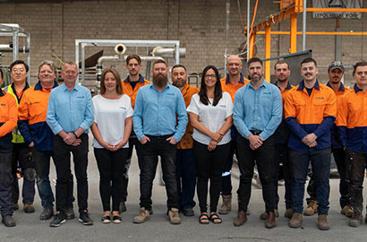 A group of workers stands together in a warehouse. Some wear blue shirts, while others wear orange and navy uniforms. They are facing the camera with a background of industrial equipment.