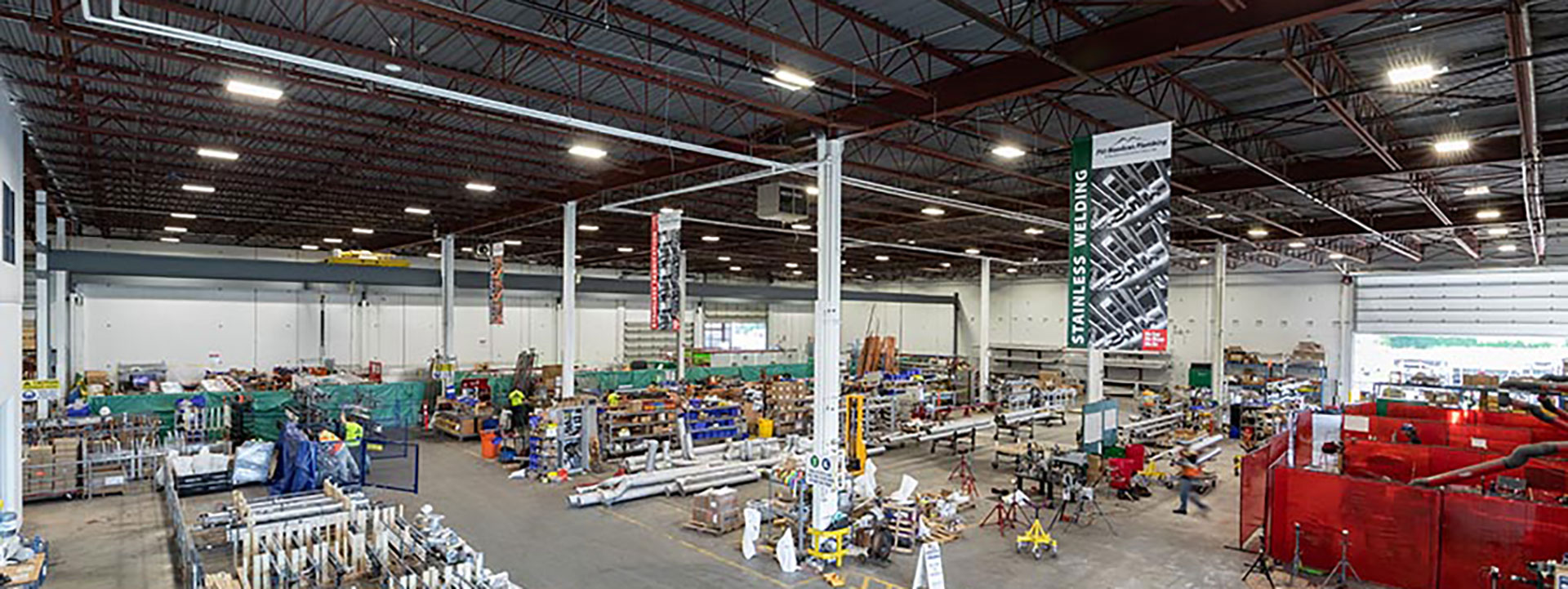 A large, spacious warehouse with various industrial equipment and materials. Workers in safety gear are scattered throughout the area.