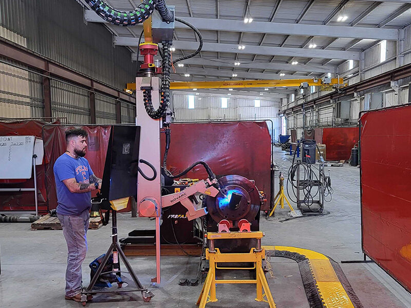 A man operates a robotic welding machine in a large industrial warehouse. The machine is positioned near a protective red screen, with various equipment and tools visible in the background. The warehouse has a high ceiling and bright lighting.