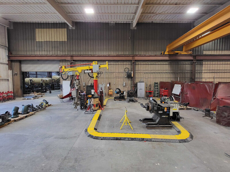 A spacious industrial workshop featuring various machinery and equipment, including a yellow overhead crane and several workstations. The floor is marked with yellow safety lines, and the area is well-lit with overhead lights.