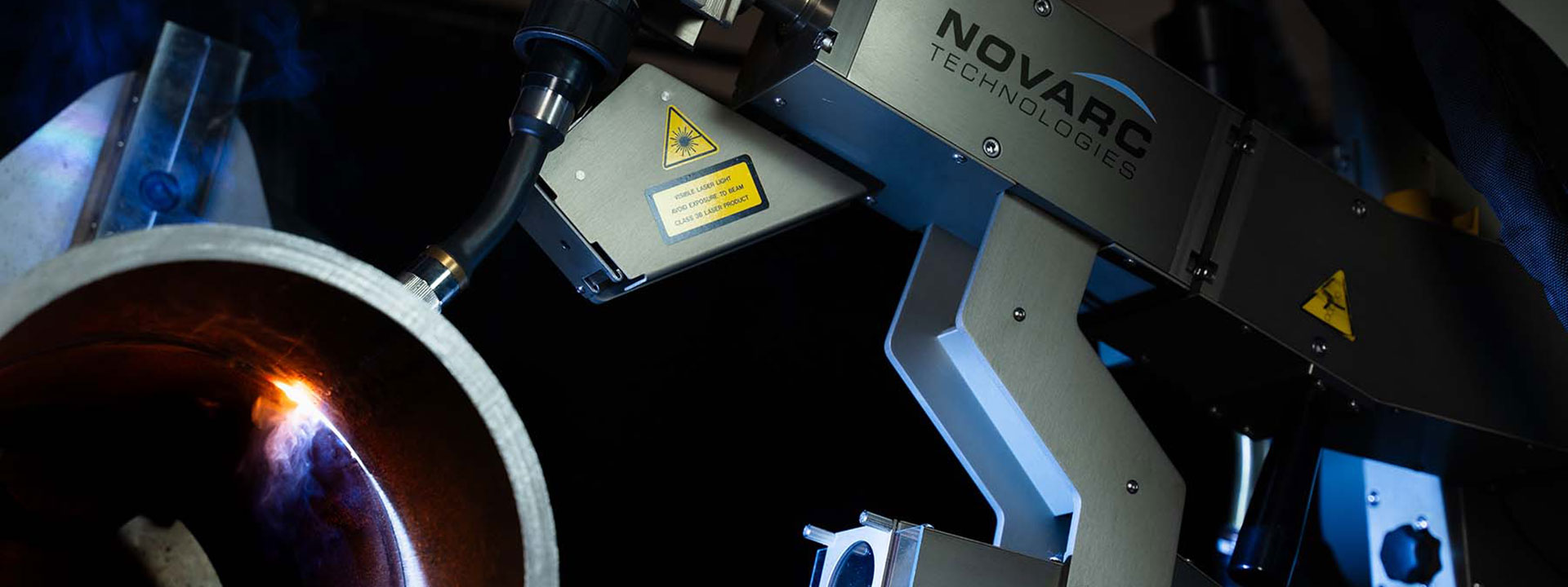 A high-tech industrial welding machine by Novarc Technologies is in action, with sparks and a bluish glow indicating active welding on a curved metal surface.