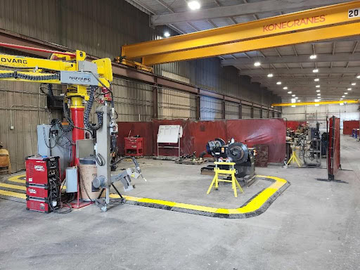 A large industrial workshop with a yellow overhead crane labeled "Konecranes." Various equipment, including a robotic arm and welding machines, are arranged in the space. Red divider curtains section off areas within the facility.