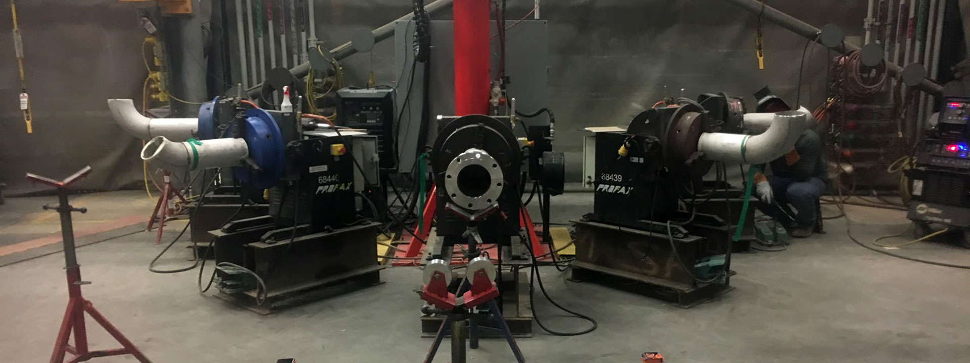 A room with industrial machinery, including large pipes and blowers, positioned on metal platforms. A person works in the background amidst wires and equipment.