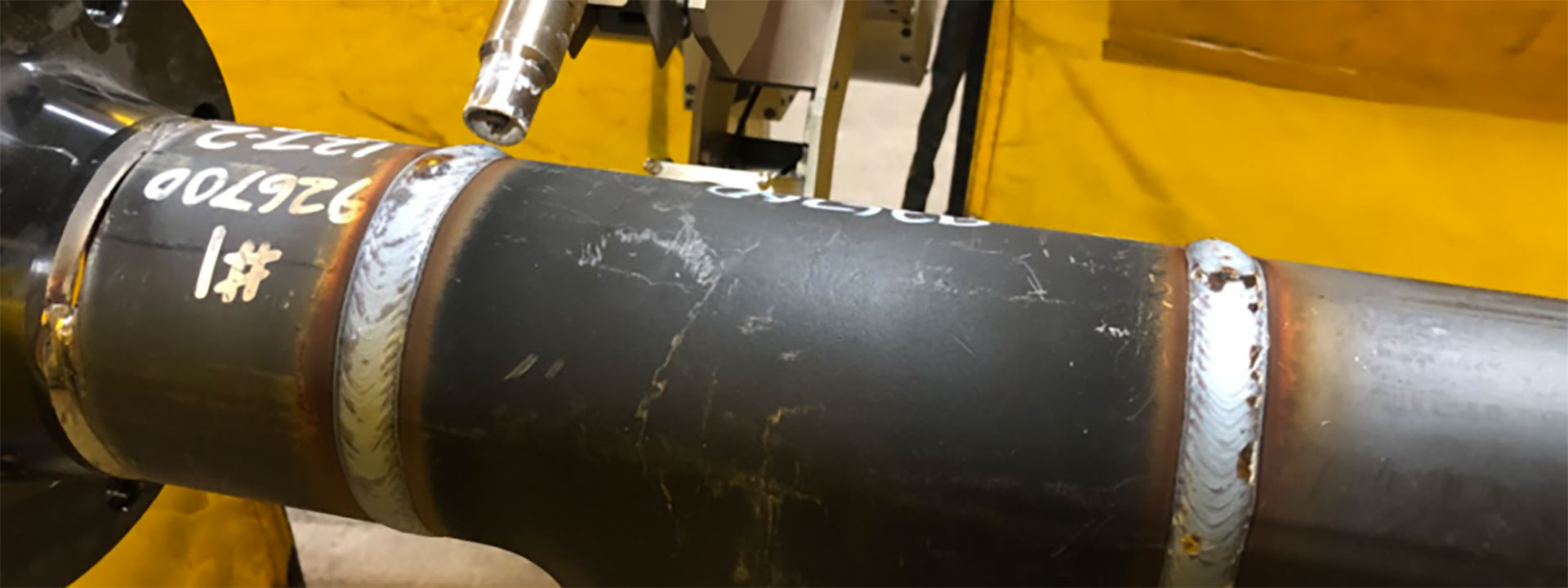 A close-up of a welded metal pipe section in an industrial setting. The pipe has several visible weld seams and white markings with numbers.