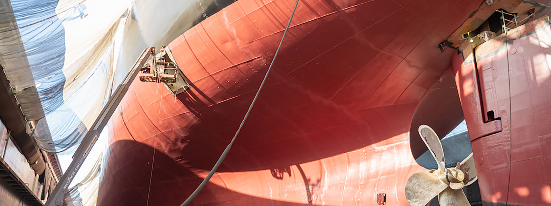 View of a ship's massive red hull in dry dock, with sunlight casting shadows. Visible is a large propeller at the bottom right, and a rope or cable hanging down.