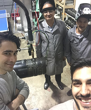 Four people in lab coats and safety gear stand smiling around a piece of industrial equipment in a workshop. Two are wearing protective eyewear and helmets.