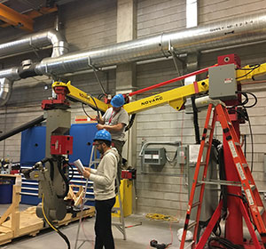 Two people wearing blue hard hats work in an industrial setting with machinery and tools. One person stands on a ladder adjusting equipment, while the other reads from a paper