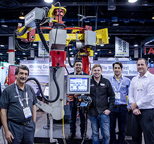 Five people stand around a large industrial welding robot at a trade show. They are posing and smiling for the photo, with booths and banners visible in the background.