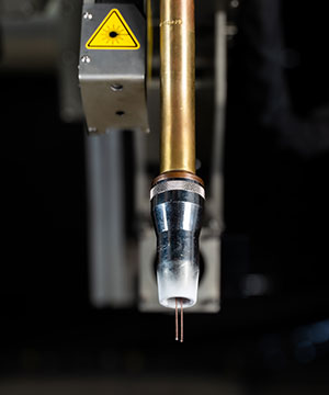 Close-up of a metal laser cutting nozzle with a warning label on the machinery above it, set against a blurred industrial background.