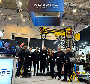 A group of people wearing black outfits stand under a large sign reading "Novarc Technologies" at an exhibition booth. In the background, there's industrial equipment displayed. The booth has bright lighting and a modern setup.