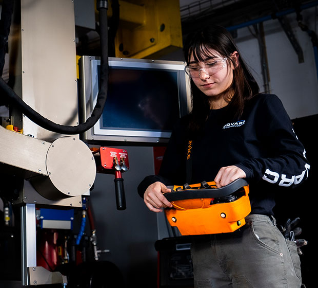 A person in safety glasses operates machinery using an orange control device. The setting is industrial, with metal equipment and monitors in the background.