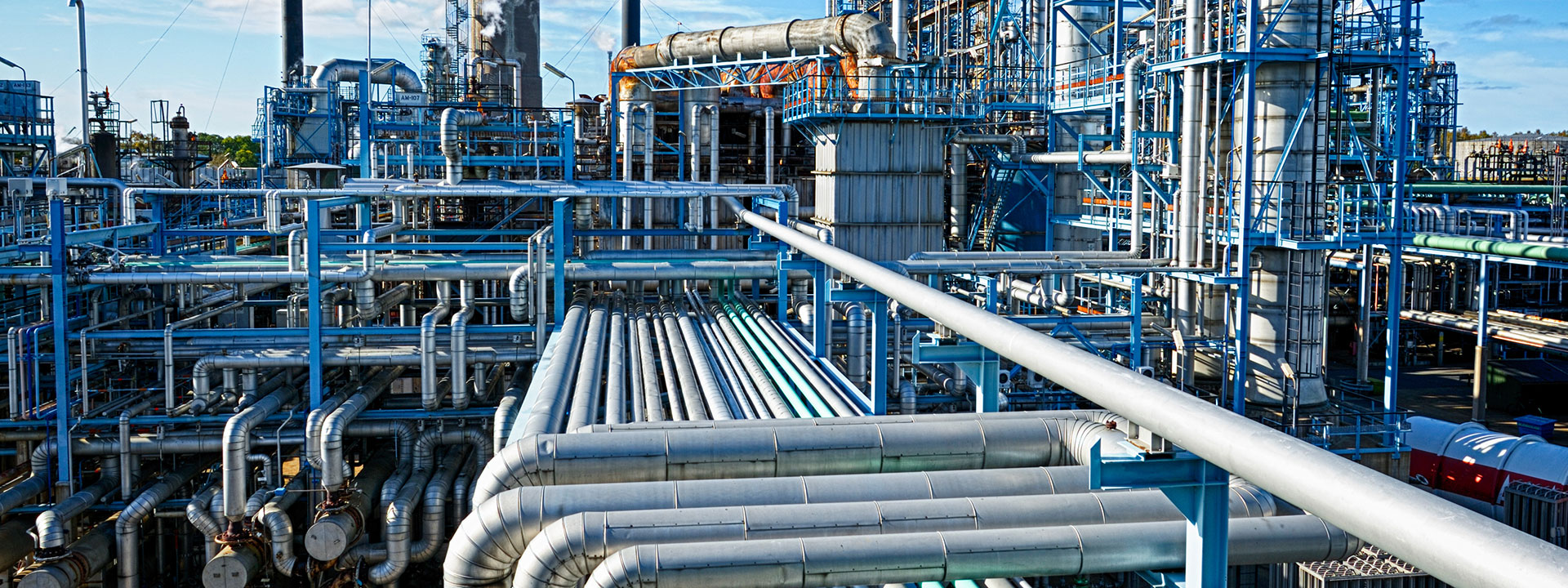 Industrial scene featuring a complex network of metal pipes and structures at a large manufacturing plant. The sky is clear and blue, highlighting the intricate engineering of the facility.