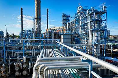 An industrial facility with complex metal structures and pipelines under a clear blue sky. Tall chimneys and intricate networks of pipes dominate the scene, suggesting a large-scale manufacturing or processing plant.