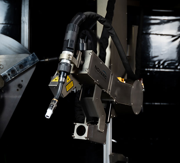 A robotic welding arm with the label "Novarc" attached to a metal stand in a dark industrial setting. The arm is equipped with various components, including a nozzle and cables,