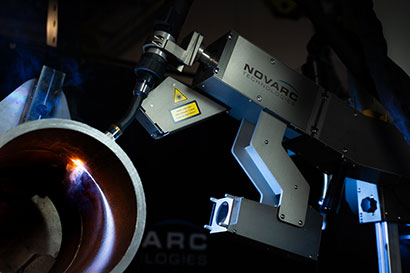 A robotic arm with the label "NOVARC" welds the edge of a large metal pipe, emitting sparks and a small amount of smoke.