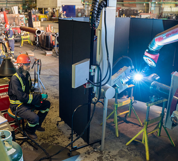 Factory worker in safety gear sits near a robotic welding machine, which emits bright sparks on a metal pipe.