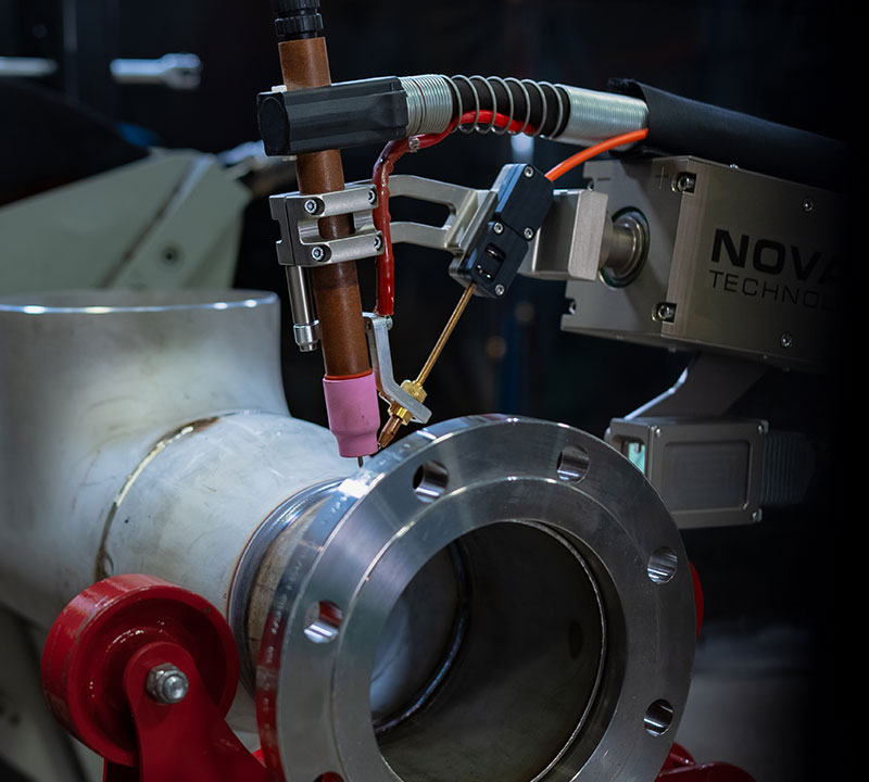 Industrial robotic arm performing precise welding on a metal pipe. The setup includes various attachments and wires for automation