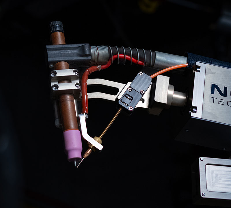 Close-up of a high-tech welding apparatus with a copper nozzle and attached cables. The equipment is set against a dark background.