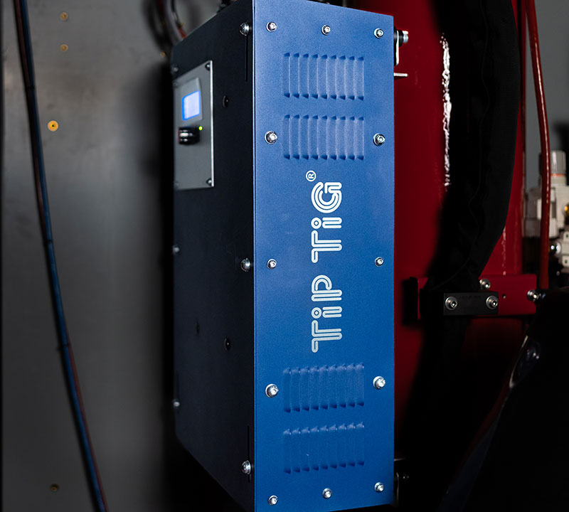 Close-up of a blue TIP TIG welding machine mounted on a dark wall. The device features visible screws and a control display screen.