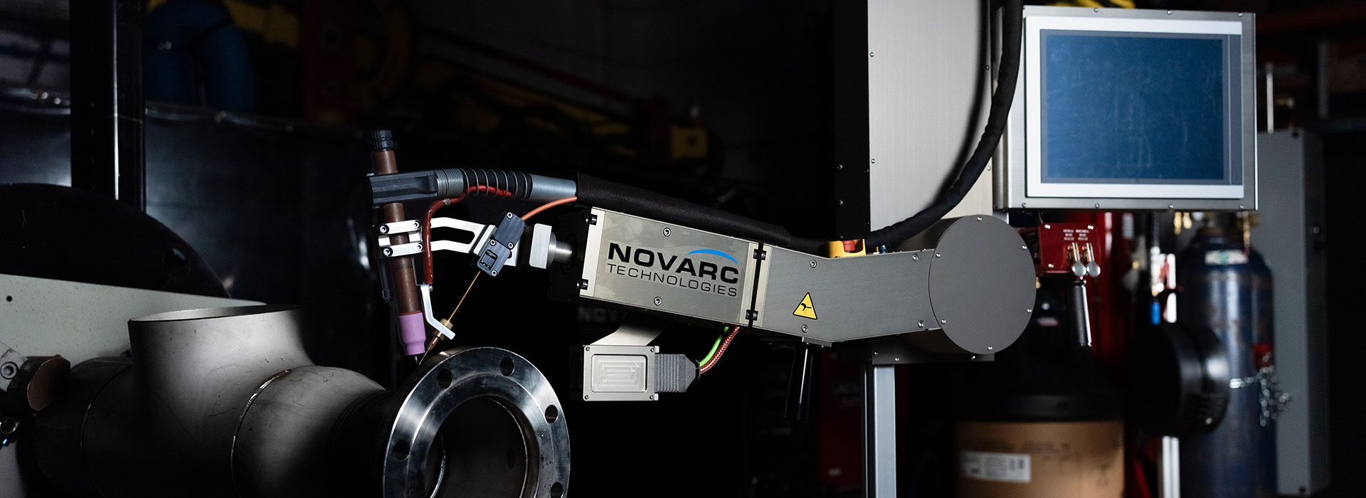 A robotic arm by Novarc Technologies is positioned near industrial piping equipment, with a screen in the background.