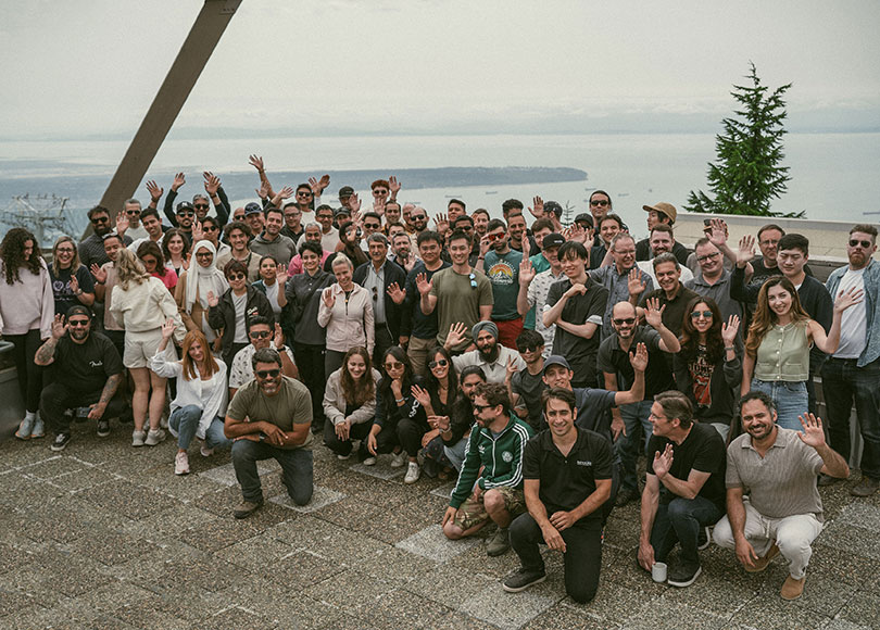 A large group of people, casually dressed, gather outdoors on a terrace with a scenic view of the sea and distant islands. They are waving and smiling at the camera. Trees and a cloudy sky are visible in the background.