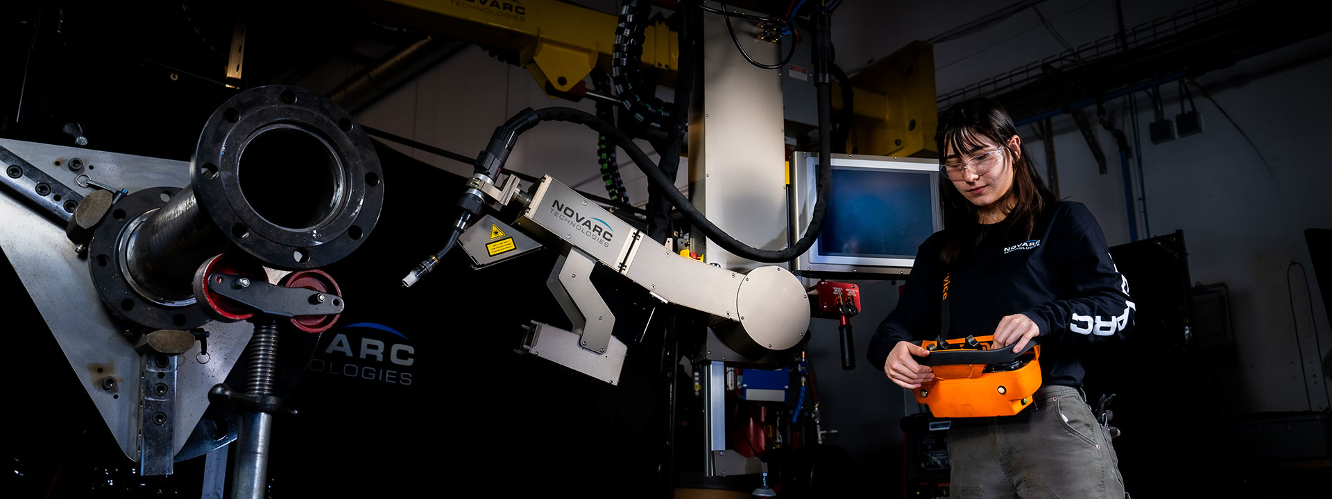 A woman operates a robotic arm in an industrial setting. She holds a control device and wears safety glasses. The machinery is labeled "Novarc Technologies," with various cables and components visible in the background.