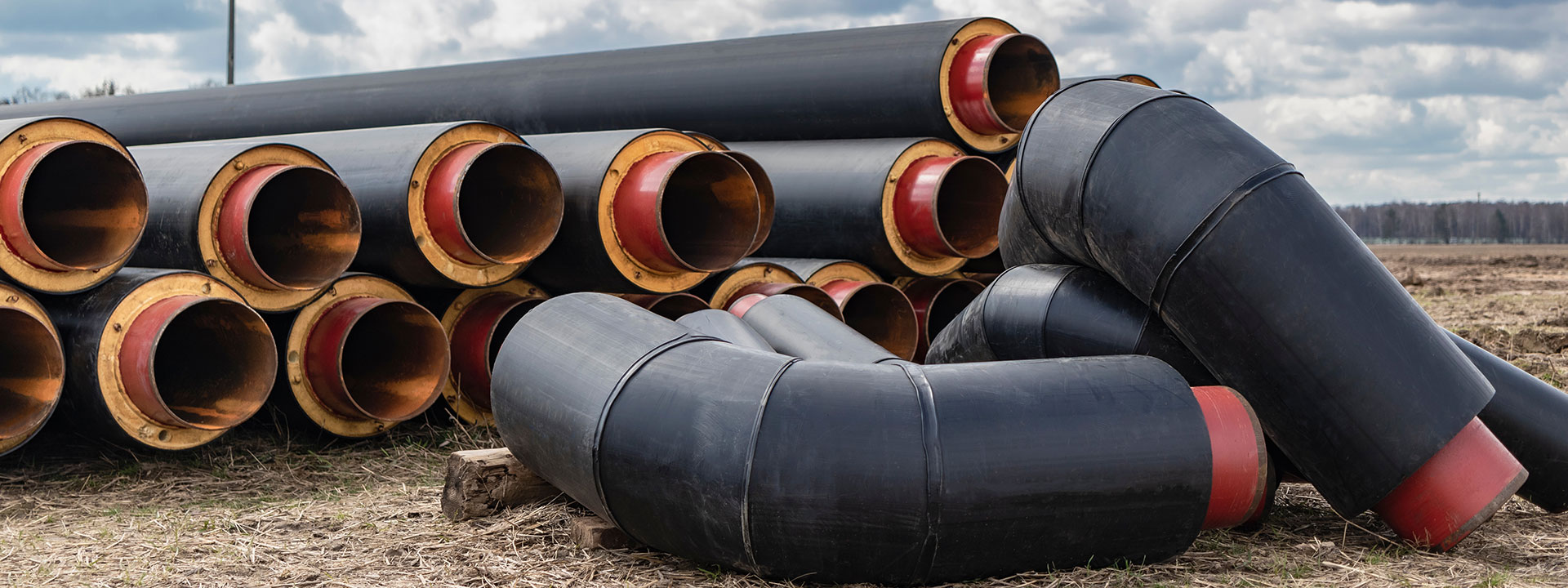 Stacks of large black metal pipes with red linings are arranged on dry grass. Several curved pipe sections are placed in the foreground.