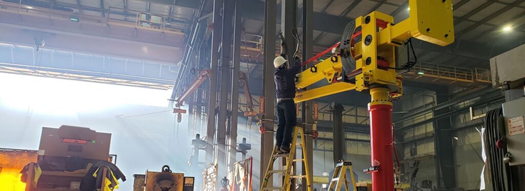 A worker in a hard hat stands on a ladder inside a large industrial factory, adjusting a piece of heavy machinery with yellow and red components.