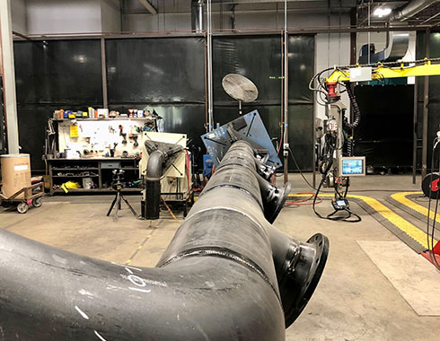 A large industrial workshop showing a long black metal pipe with several joints and flanges on the floor.