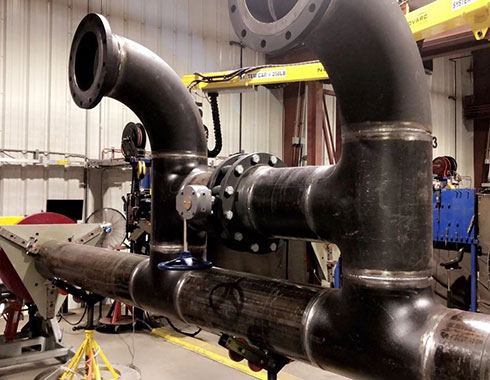 A large, industrial piping system with multiple welded joints is set up in a workshop. The pipes are connected at right angles, and various clamps and supports are holding them in place.