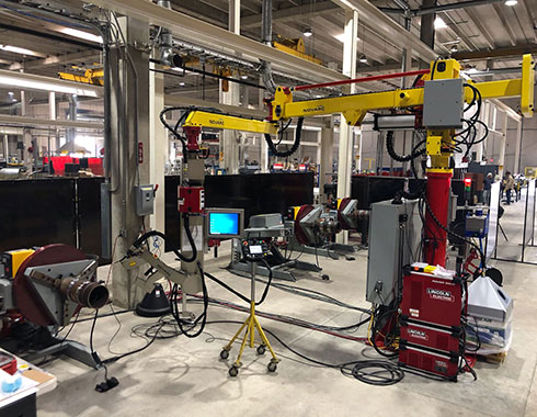 A large industrial workshop with machinery, robotic arms, and control panels. There's a computer screen mounted on a stand in the center.